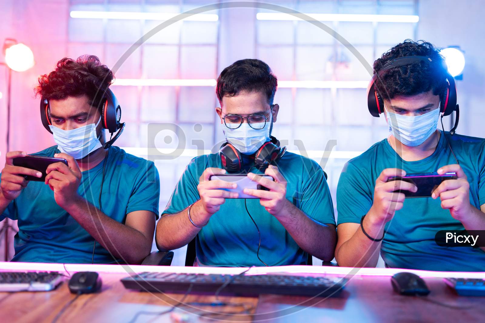 Group Of Gamers With Medical Face Mask Playing Video Game On Mobile Phone At Esports League Or Tournament During Coronavirus Or Covid-19 With Safety Measures.