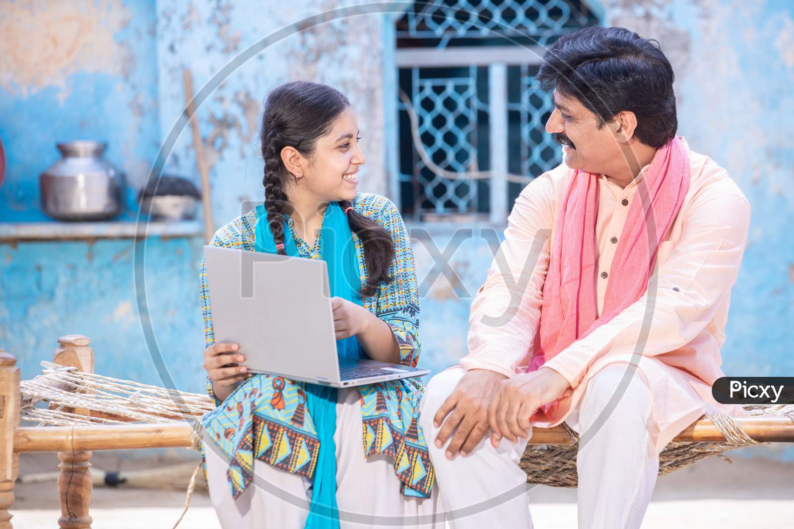 Cheerful Indian Father And Young Daughter Using Laptop Looking At Each Other While Sitting On Traditional Wooden Bed Outside Their House, Happy Rural Family, Web Surfing, Technology Concept.