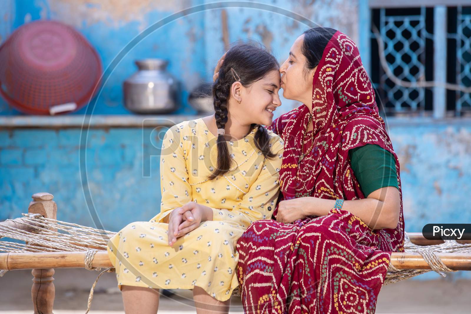 Young Rural Indian Mother Kiss Smiling Little Daughter In Forehead, Caring Loving Traditional Mom In Red Sari Supports And Motivates Small Preschooler Girl Child,Sitting On Bed At Village Home.