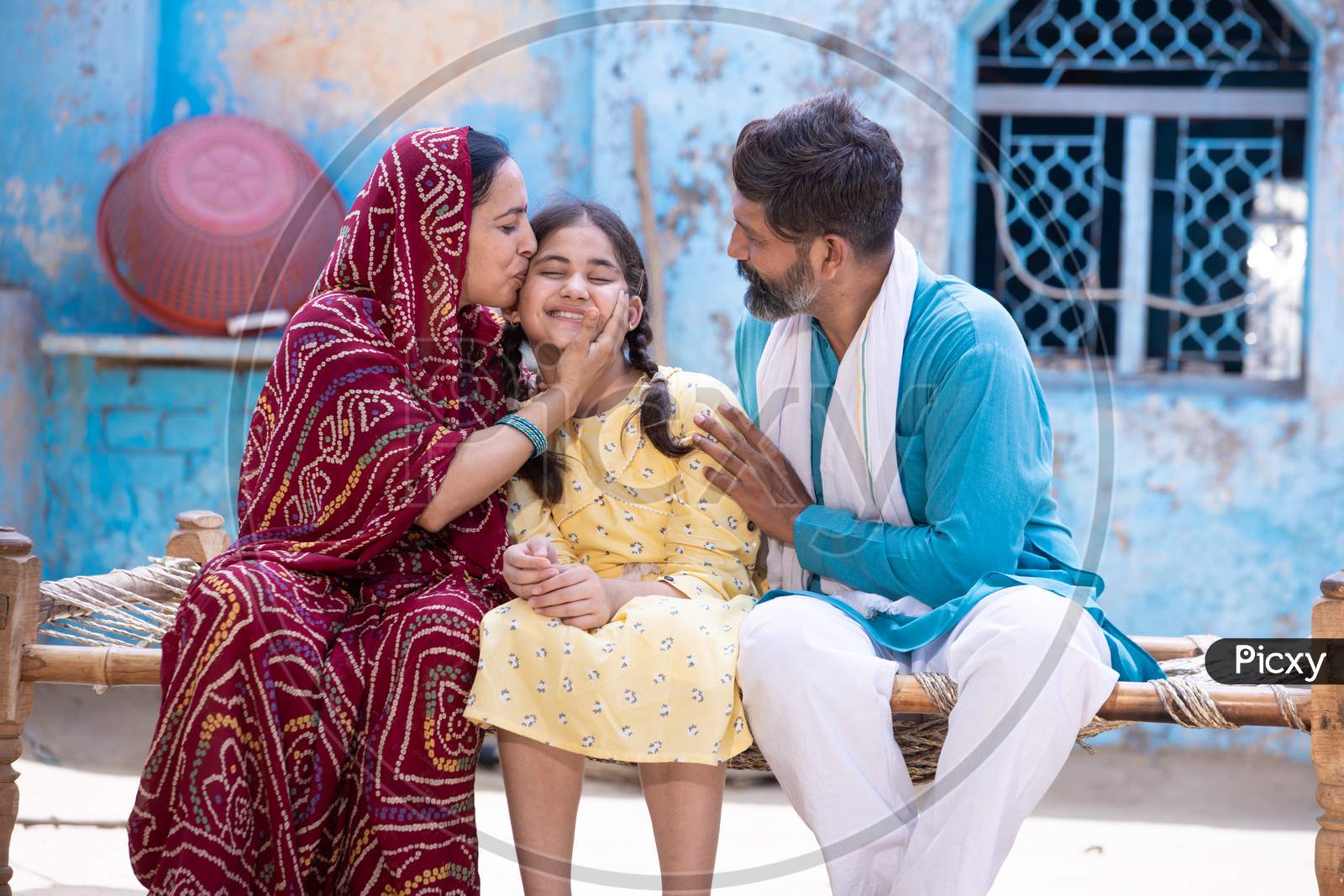 Happy Rural Indian Family Smiling While Sitting On Traditional Bed At Village Home, Mother Is Sari Hug And Kiss Her Cute Little Daughter And Father Looking At Her. Parent Love Their Child.