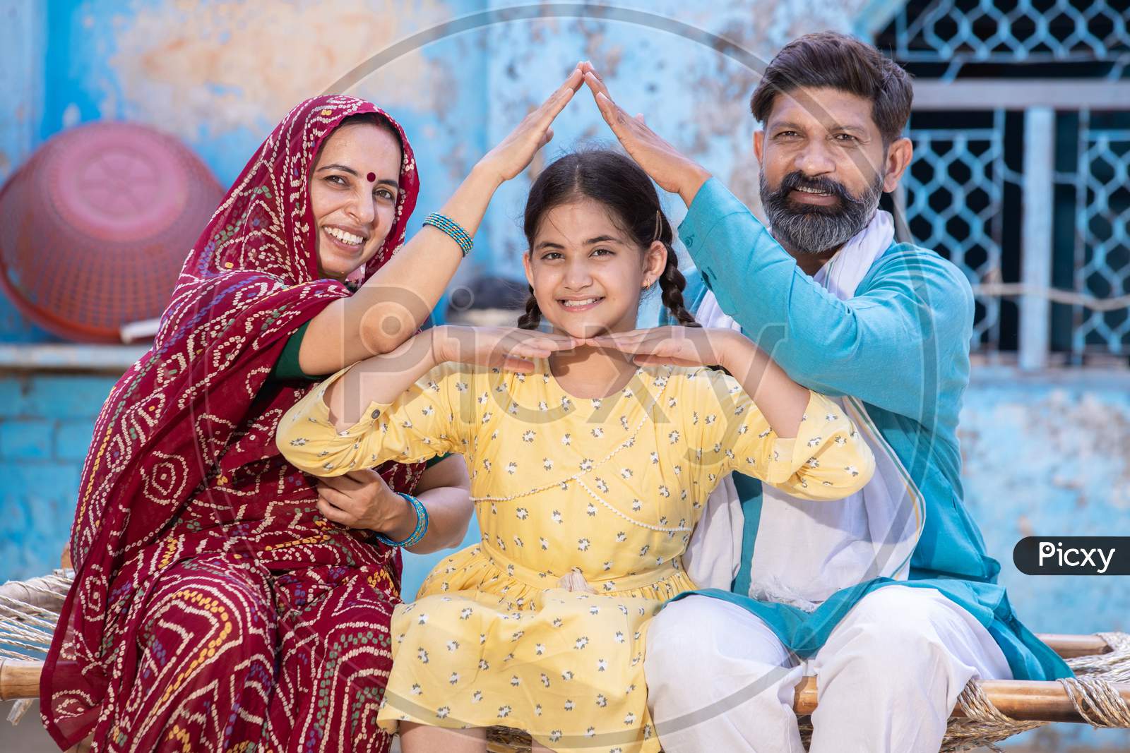 Portrait Of Smiling Happy Rural Indian Family Forming House Roof With Their Hands At Village Home, Parents Care For Child Protection, Father And Mother With Daughter, India Home Loan And Insurance Concept.