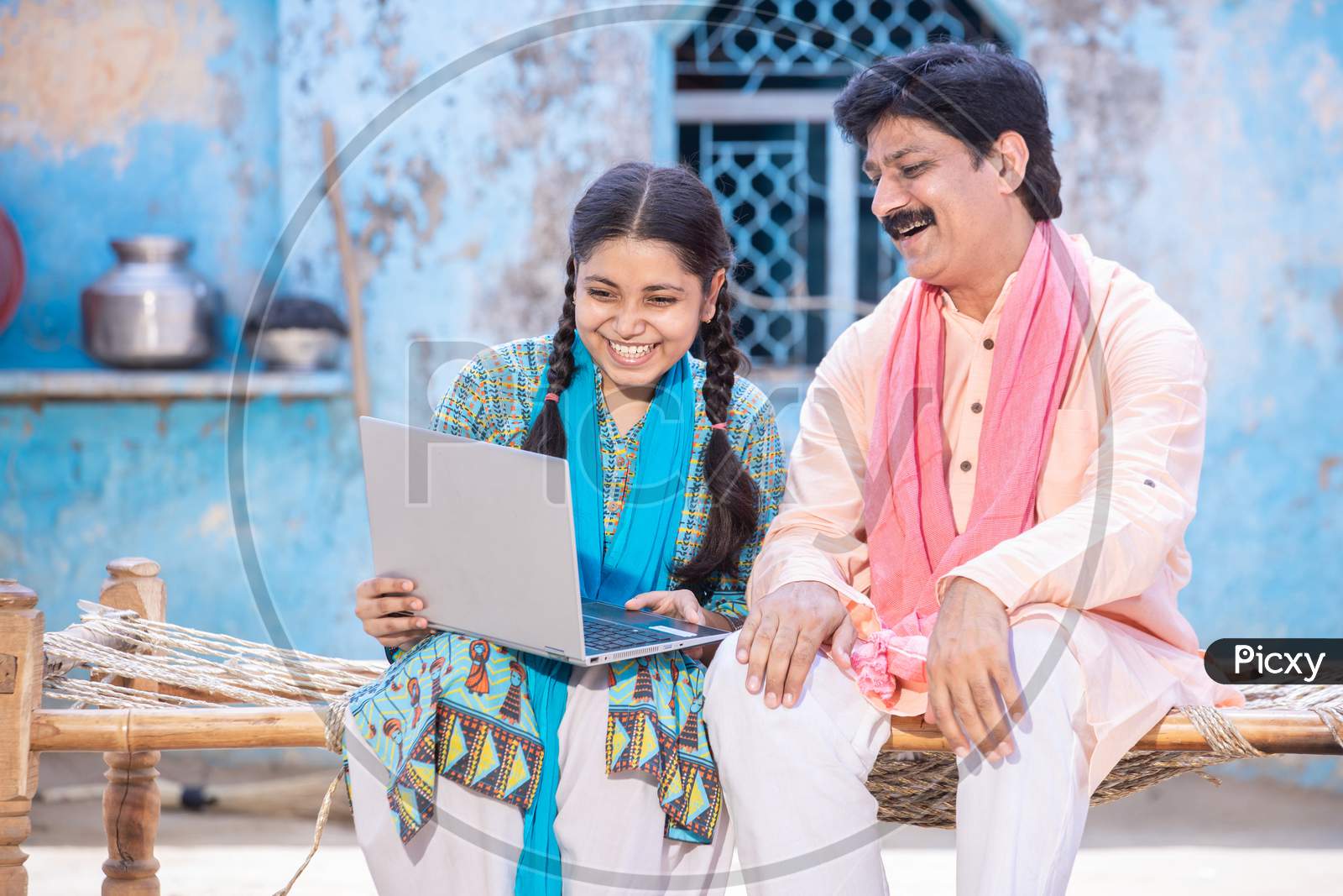 Pleasant Indian Father And Young Daughter Using Laptop While Sitting On Traditional Wooden Bed Outside Their House, Happy Rural Family Looking At Computer Screen. Web Surfing. Technology Concept.