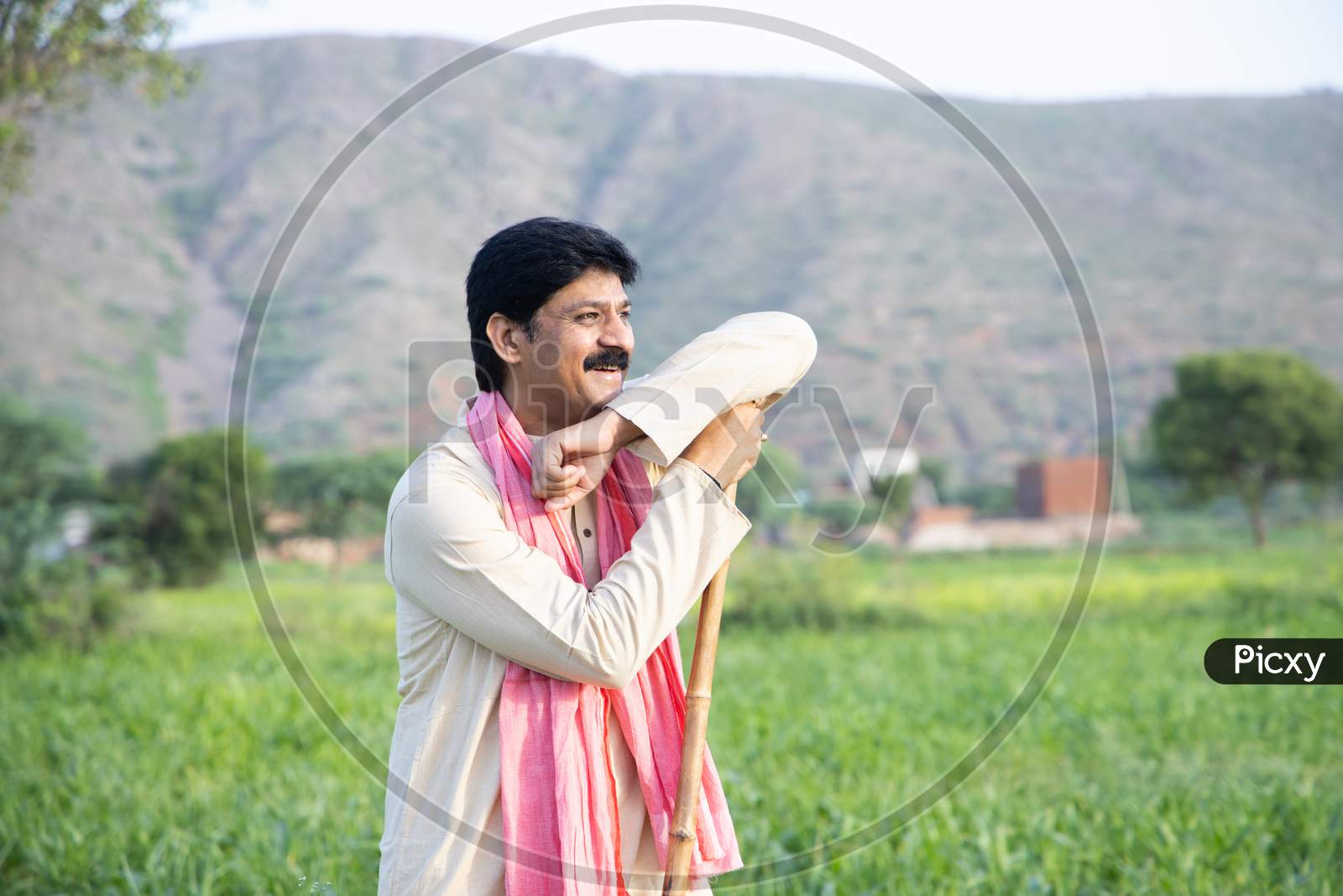 Happy Indian Farmer Holding Wood Stick In Hand Standing In Agriculture Field Wearing Traditional Kurta Dress, Smiling Man With Mustache And Black Hair. Rural India Concept. Copy Space.