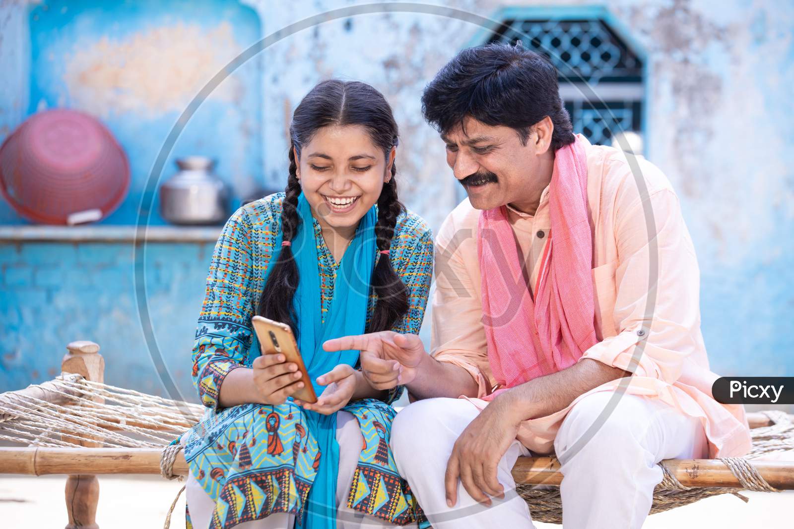 Happy Indian Father And Young Daughter Using Smartphone While Sitting On Traditional Bed Outside Their House, Happy Rural Family, Man Point Finger At Mobile Screen. Video Call, Learning Technology