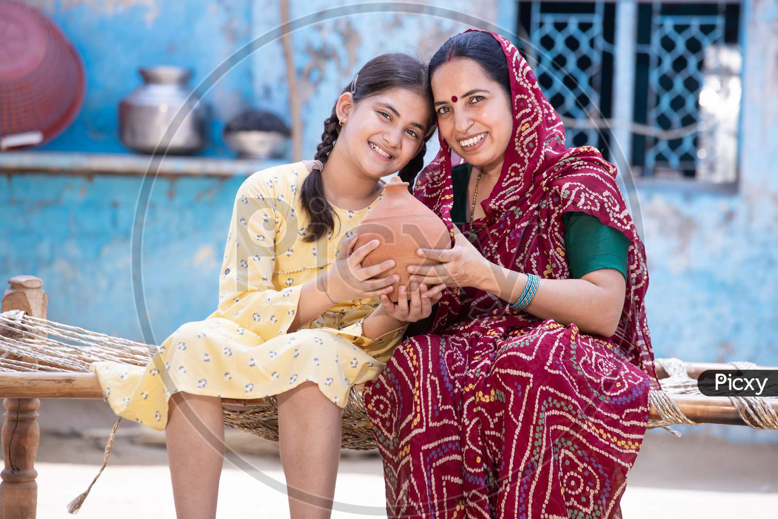 Portrait Of Happy Rural Indian Mother And Adorable Daughter Holding Piggy Bank While Sitting On Traditional Bed At Village Home, Cute Little Girl Collect Money, Saving And Investment Concept.