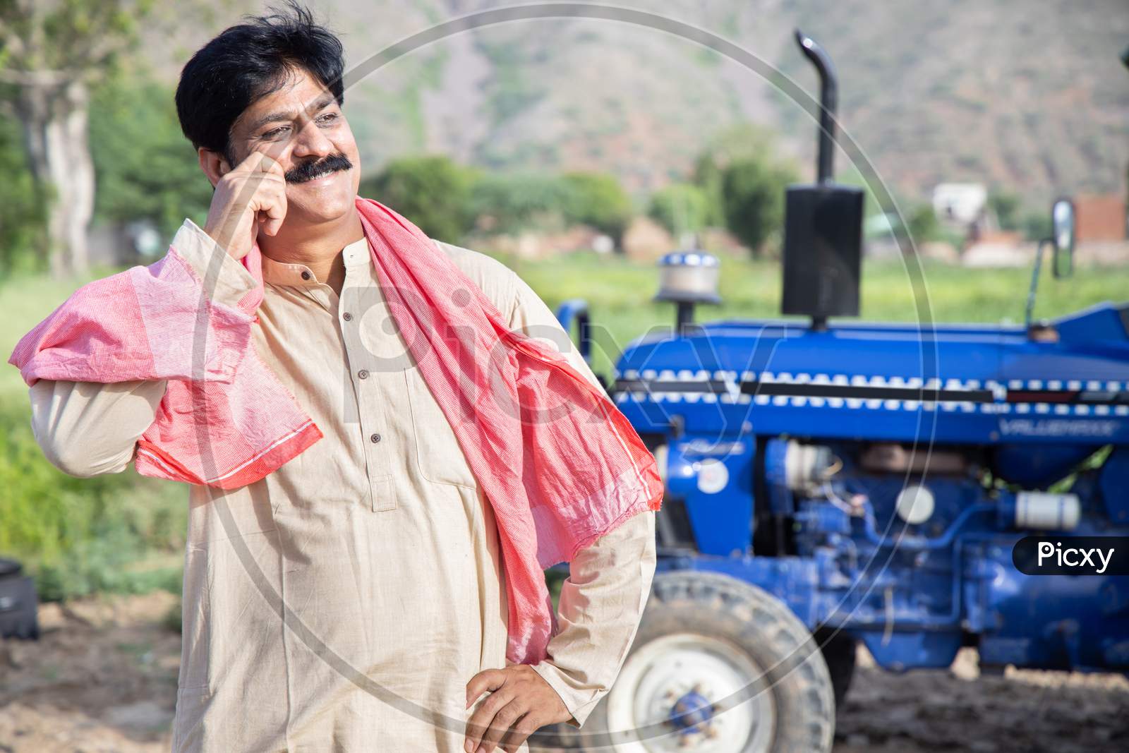 Young Happy Indian Farmer Touching His Mustache Standing At Agriculture Field With Blue Tractor Behind Him. Man Wearing Traditional Kurta Feel Proud. Rural India Concept.