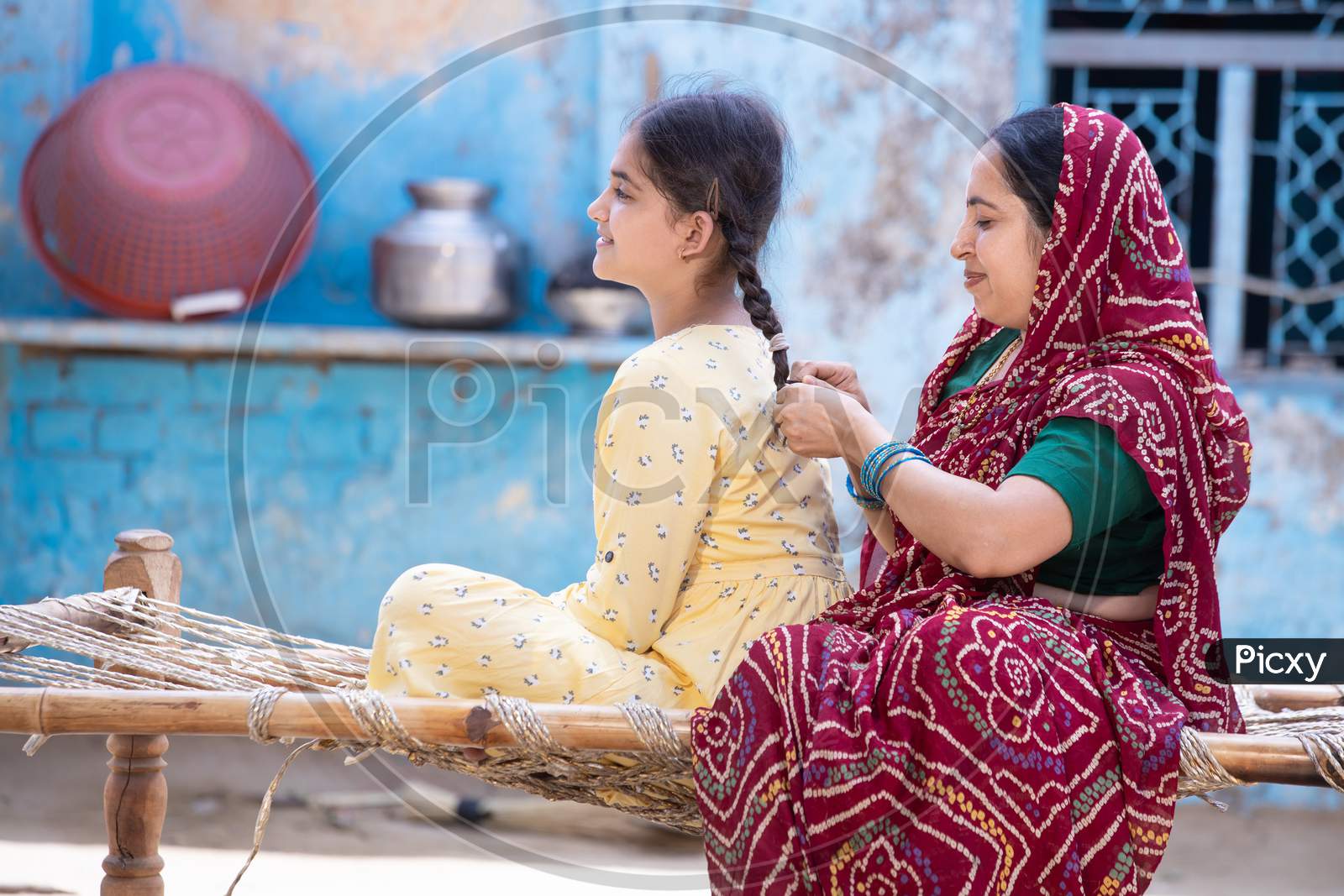 Rural Indian Mother Braids Hair Of Her Young Adorable Daughter While Sitting On Traditional Bed At Village Home. Cute Preschool Kid With Mum Spend Together, Woman In Red Sari And Girl In Yellow Dress
