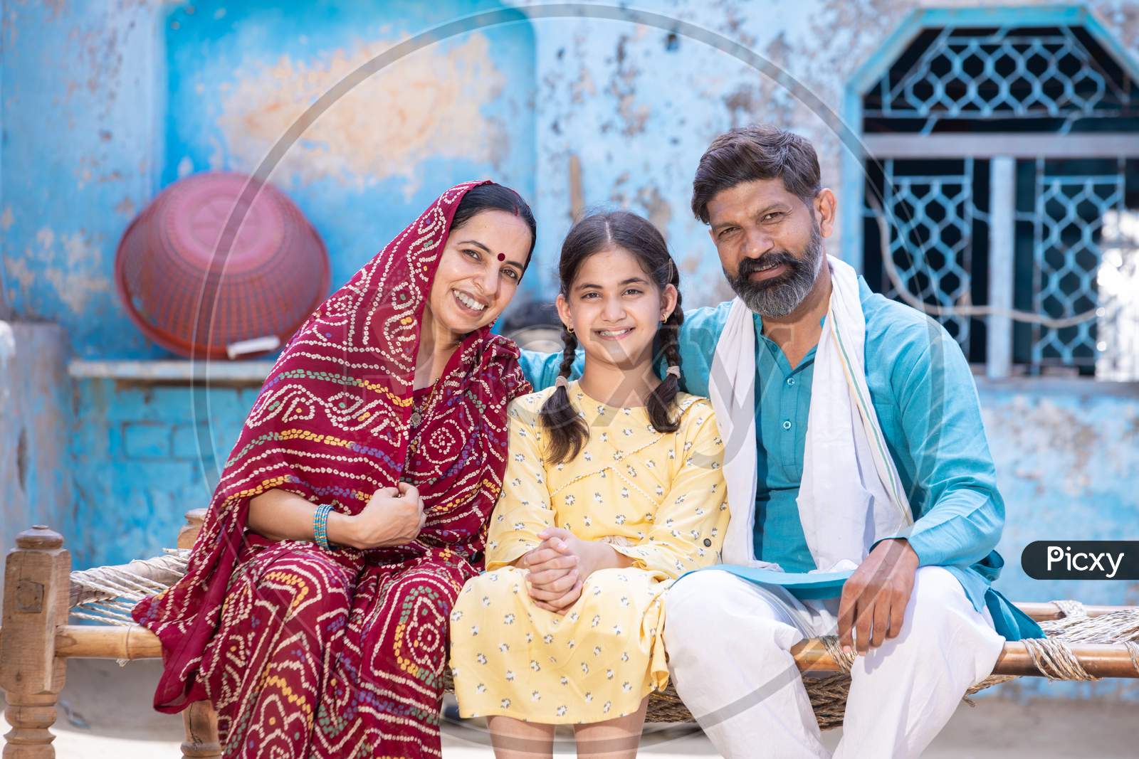 Portrait Of Happy Rural Indian Family Looking At Camera While Sitting On Traditional Bed At Village Home,Young Husband And Wife With Cute Little Daughter. One Child Policy Concept