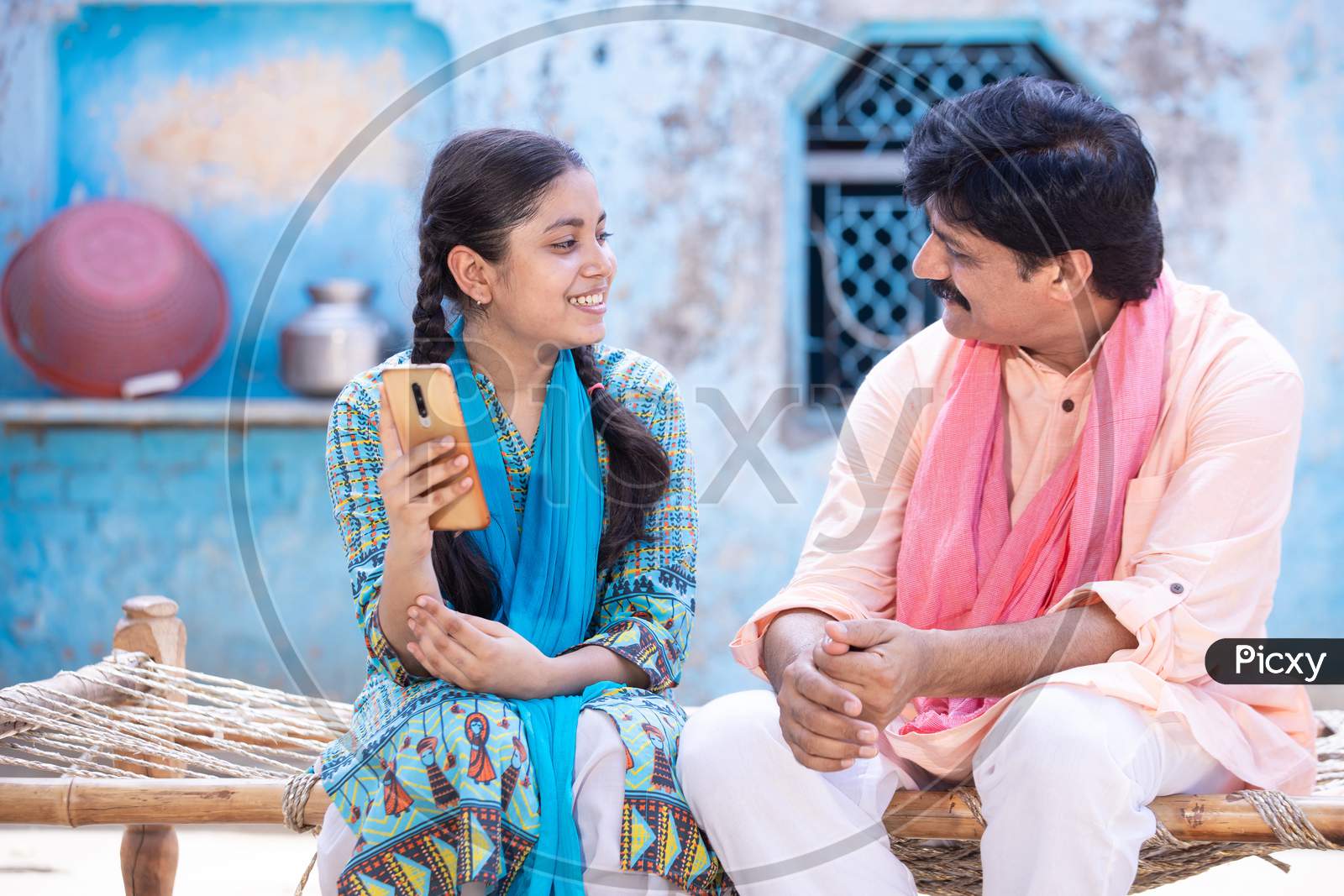 Cheerful Indian Father And Young Daughter Using Smartphone Looking At Each Other While Sitting On Traditional Wooden Bed Outside Their House, Happy Rural Family, Technology Concept.