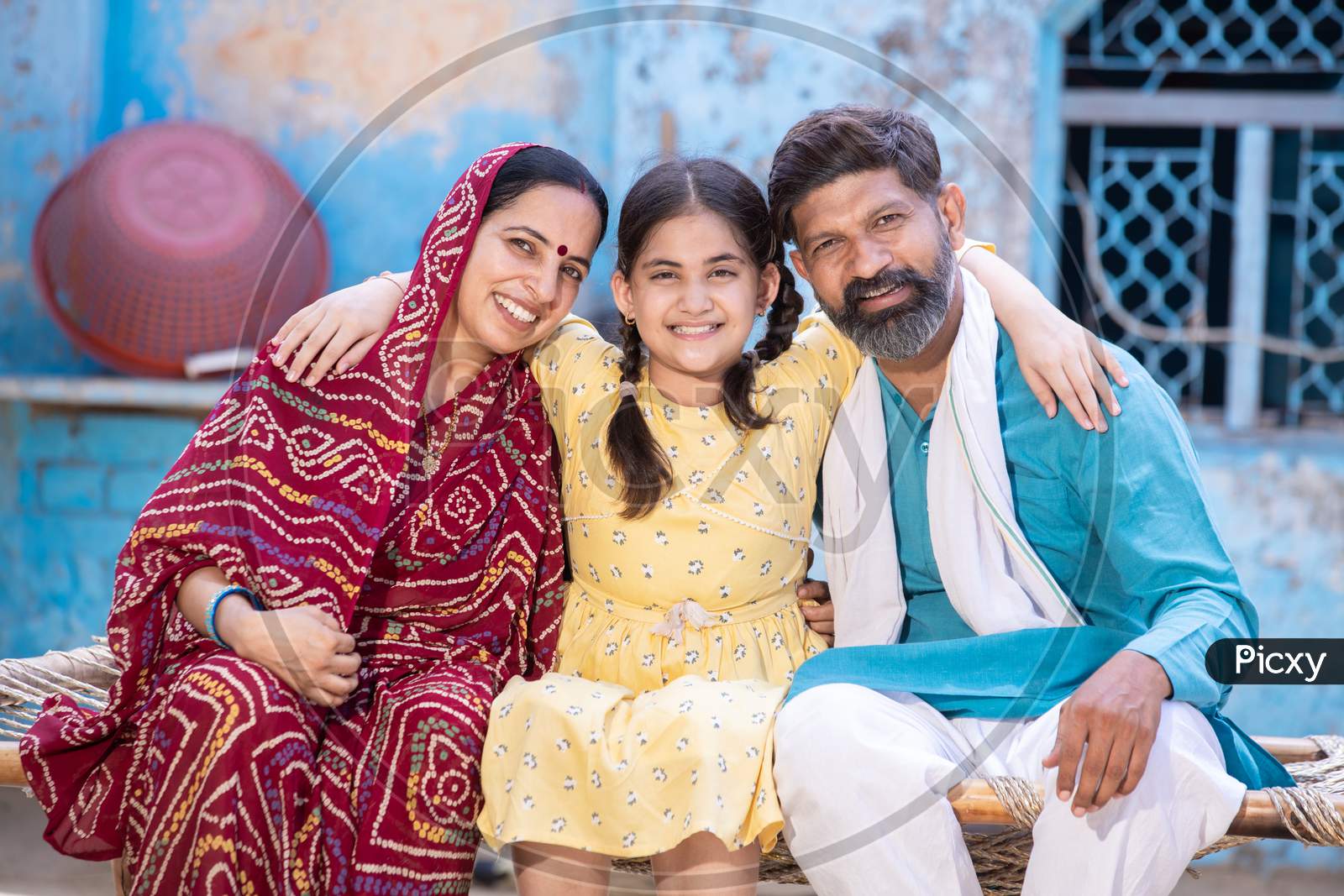 Portrait Of Happy Rural Indian Family In Looking At Camera While Sitting On Traditional Bed At Village Home, Cute Adorable Little Daughter Hug Her Father And Mother. India Parent With Child Smiling