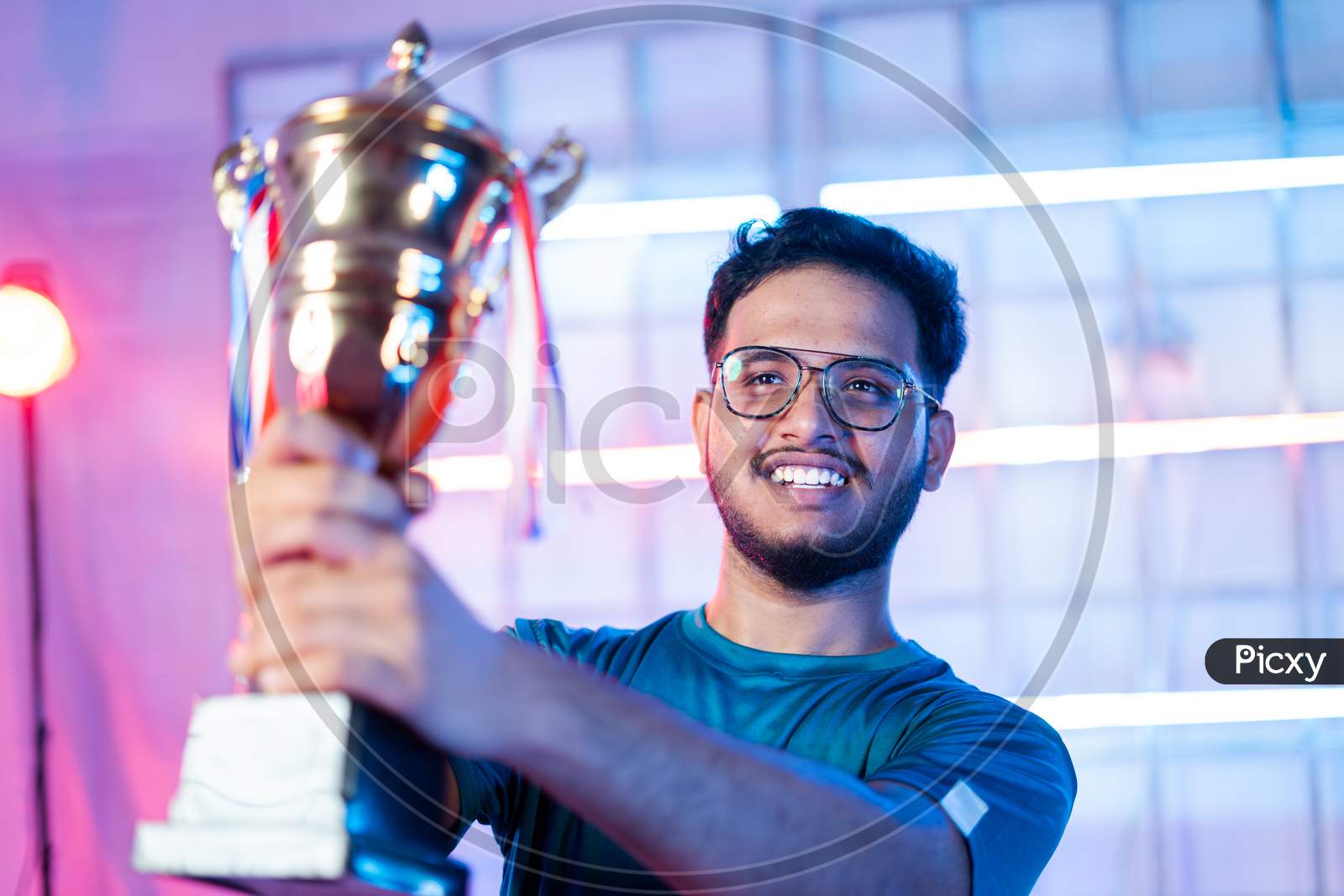 Cheerful Happy Smiling Gamer With Headphones Celebrating Byholding Winning Trophy At Esports Gaming Tournament Stage