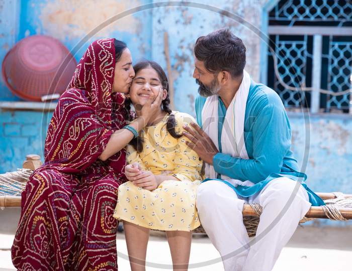 Happy Rural Indian Family Smiling While Sitting On Traditional Bed At Village Home, Mother Is Sari Hug And Kiss Her Cute Little Daughter And Father Looking At Her. Parent Love Their Child.