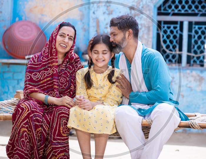 Happy Rural Indian Family Smiling While Sitting On Traditional Bed At Village Home, Father Is Kissing Her Cute Little Daughter On Head And Mother In Sari Looking At Camera Parent Support Love Child.