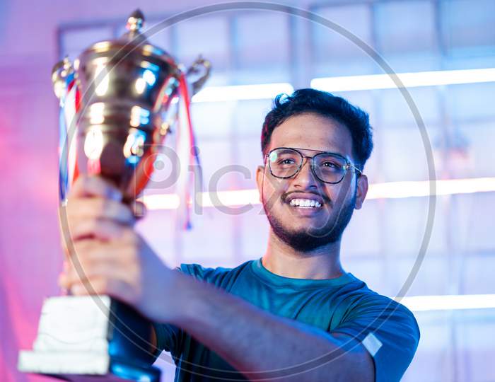 Cheerful Happy Smiling Gamer With Headphones Celebrating Byholding Winning Trophy At Esports Gaming Tournament Stage