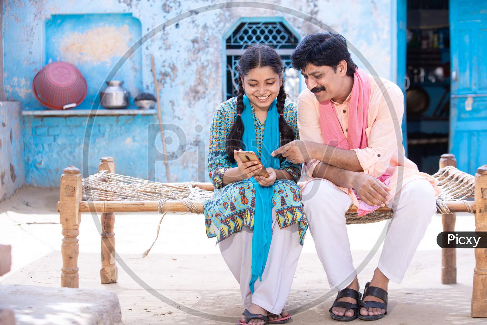 Happy Indian Father And Young Daughter Using Smartphone While Sitting On Traditional Bed Outside Their House, Happy Rural Family, Village Man Learning Mobile Phone Technology, Copy Space.