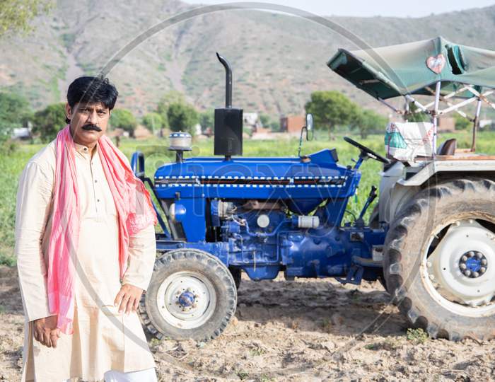 Young Happy Indian Farmer Standing Agriculture Field With Blue Tractor Behind Him. Man Posing Wearing Traditional Kurta With Mustache Looking At Camera. Rural India Concept.
