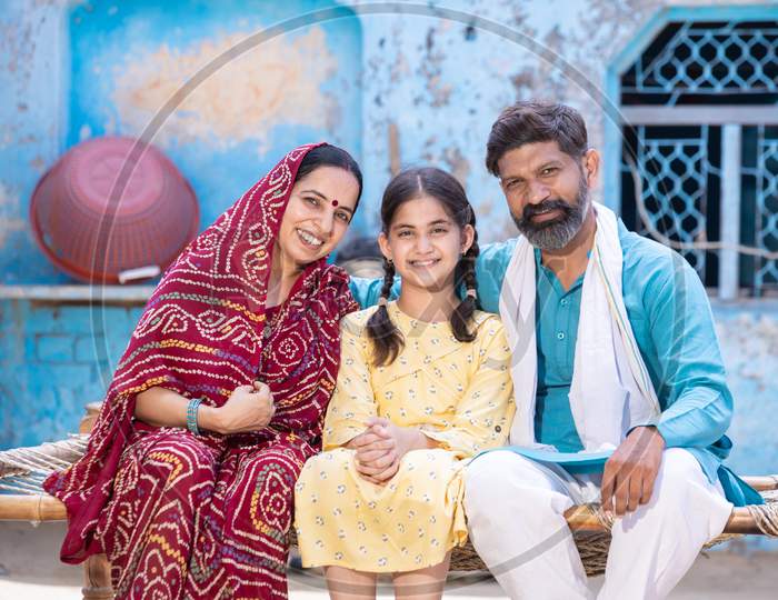 Portrait Of Happy Rural Indian Family Looking At Camera While Sitting On Traditional Bed At Village Home,Young Husband And Wife With Cute Little Daughter. One Child Policy Concept