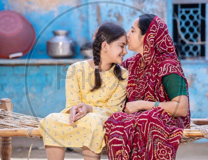 Young Rural Indian Mother Kiss Smiling Little Daughter In Forehead, Caring Loving Traditional Mom In Red Sari Supports And Motivates Small Preschooler Girl Child,Sitting On Bed At Village Home.