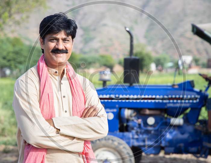 Portrait Of Young Happy Indian Farmer Standing With Blue Tractor At Agriculture Field. Man With Cross Arms Wearing Traditional Kurta Smiling Looking At Camera. Rural India Concept.