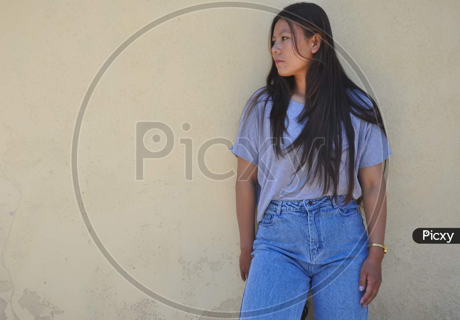 Photo of a south asian young girl with wearing gray tshirt and blue jean standing against yellow wall with looking her right side with copy space