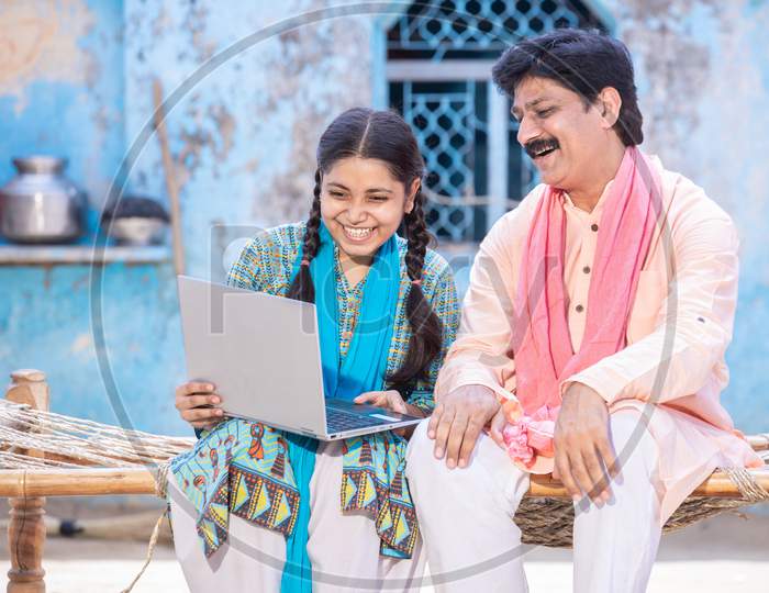 Pleasant Indian Father And Young Daughter Using Laptop While Sitting On Traditional Wooden Bed Outside Their House, Happy Rural Family Looking At Computer Screen. Web Surfing. Technology Concept.