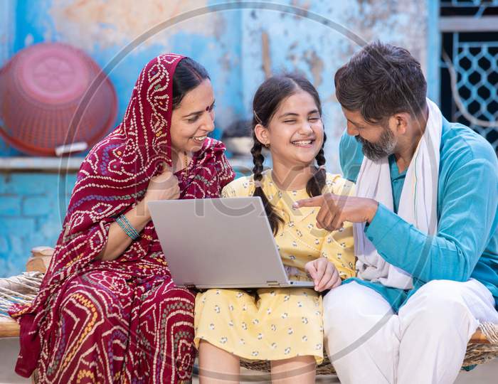 Happy Indian Little Girl Child Using Laptop While Sitting With Her Father And Mother. Village Parents Motivate Support Their Daughter For Online Education. Digital India. Man Point Finger At Computer