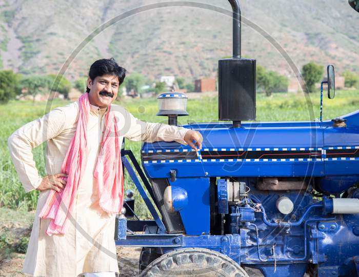 Portrait Of Young Happy Indian Farmer Standing With Blue Tractor At Agriculture Field. Man Wearing Traditional Kurta With Mustache Smiling Looking At Camera. Rural India Concept.