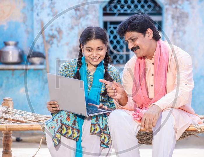 Happy Indian Father And Young Daughter Using Laptop While Sitting On Traditional Bed Outside Their House, Happy Rural Family Learning Computer, Man Pointing Towards Display, Web Surfing, Technology Concept.