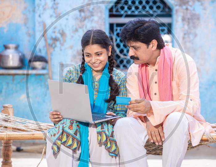 Rural Indian Father And Young Daughter Using Laptop Doing Online Payment Holding Bankcard, Stay At Home. Family Looking At Computer Screen Paying Utilities Bills Or Making Purchases On Internet.