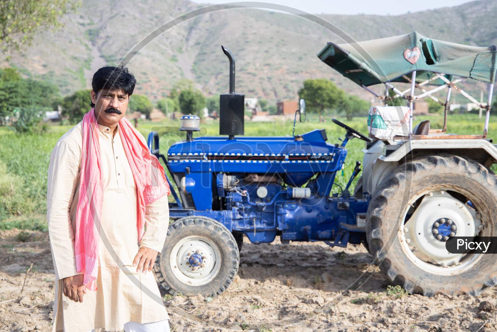 Young Happy Indian Farmer Standing Agriculture Field With Blue Tractor Behind Him. Man Posing Wearing Traditional Kurta With Mustache Looking At Camera. Rural India Concept.
