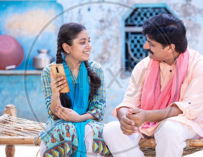 Cheerful Indian Father And Young Daughter Using Smartphone Looking At Each Other While Sitting On Traditional Wooden Bed Outside Their House, Happy Rural Family, Technology Concept.