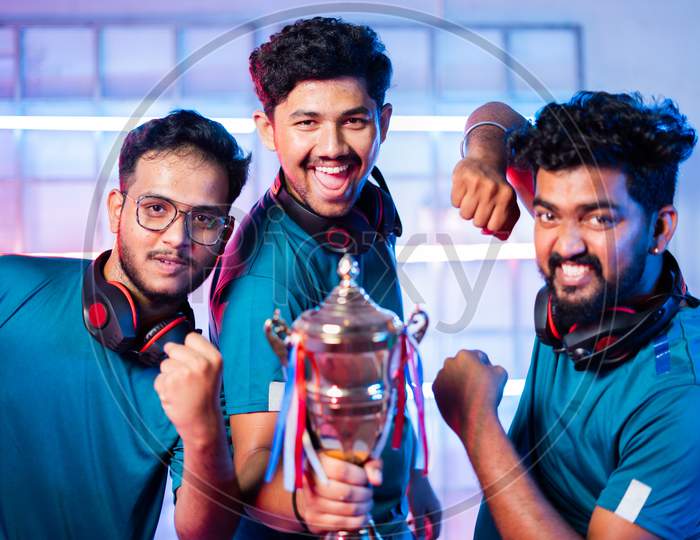 Team Of Excited Cheerful Gamers With Headphones Celebrating By Dancing And Holding Winning Trophy At Esports Gaming Tournament Stage