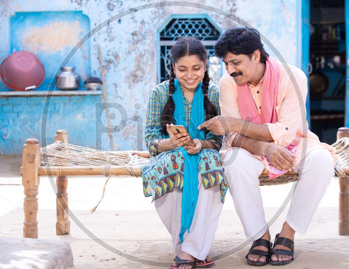 Happy Indian Father And Young Daughter Using Smartphone While Sitting On Traditional Bed Outside Their House, Happy Rural Family, Village Man Learning Mobile Phone Technology, Copy Space.