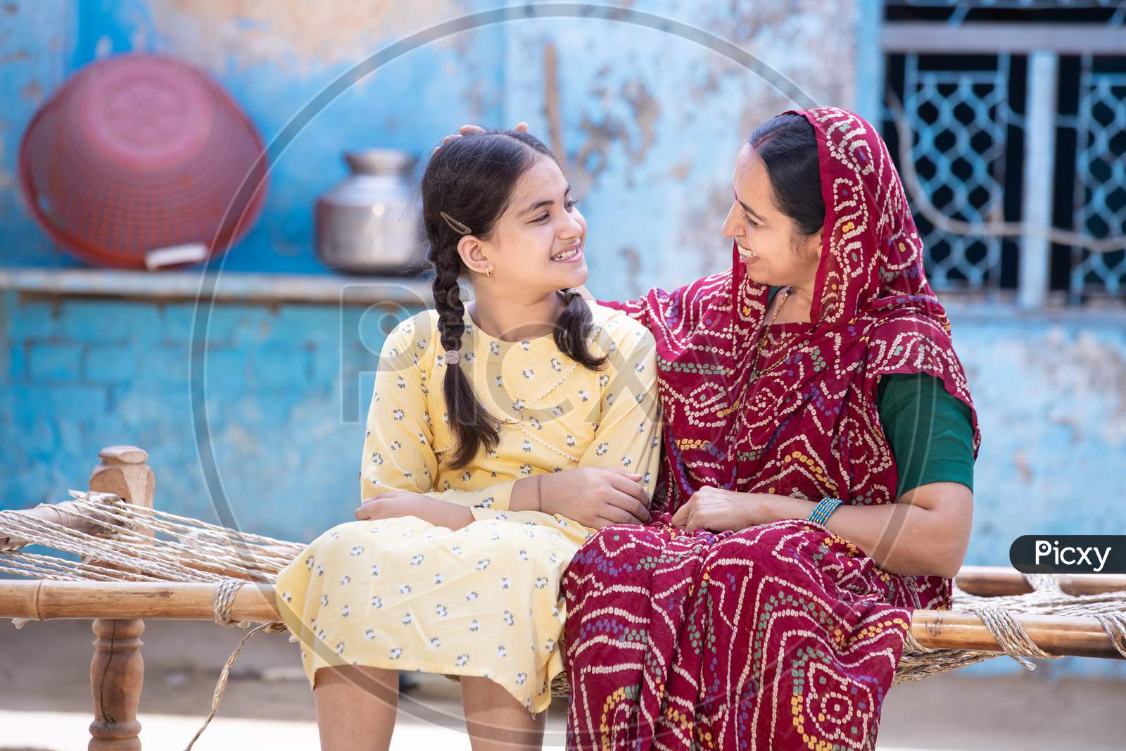 Smiling Young Indian Mother And Adorable Little Daughter Having A Good Time Together, Sitting On Traditional Bed, Cute Child Girl In Braided Hair And Mum In Red Sari Looking At Each Other.