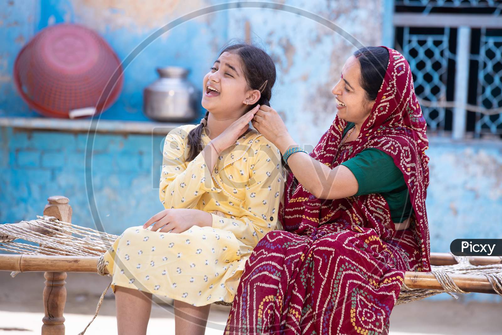 Rural Indian Mother Braids Hair Of Her Young Adorable Daughter, Cute Girl Feel Pain As Mum Getting Her Ready Sitting On Traditional Bed At Village Home. Both Spending Time Together.
