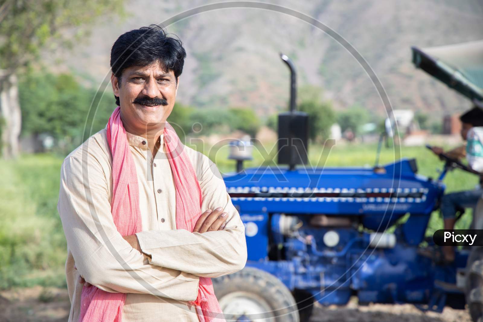 Portrait Of Young Happy Indian Farmer Standing With Blue Tractor At Agriculture Field. Man With Cross Arms Wearing Traditional Kurta Smiling Looking At Camera. Rural India Concept.