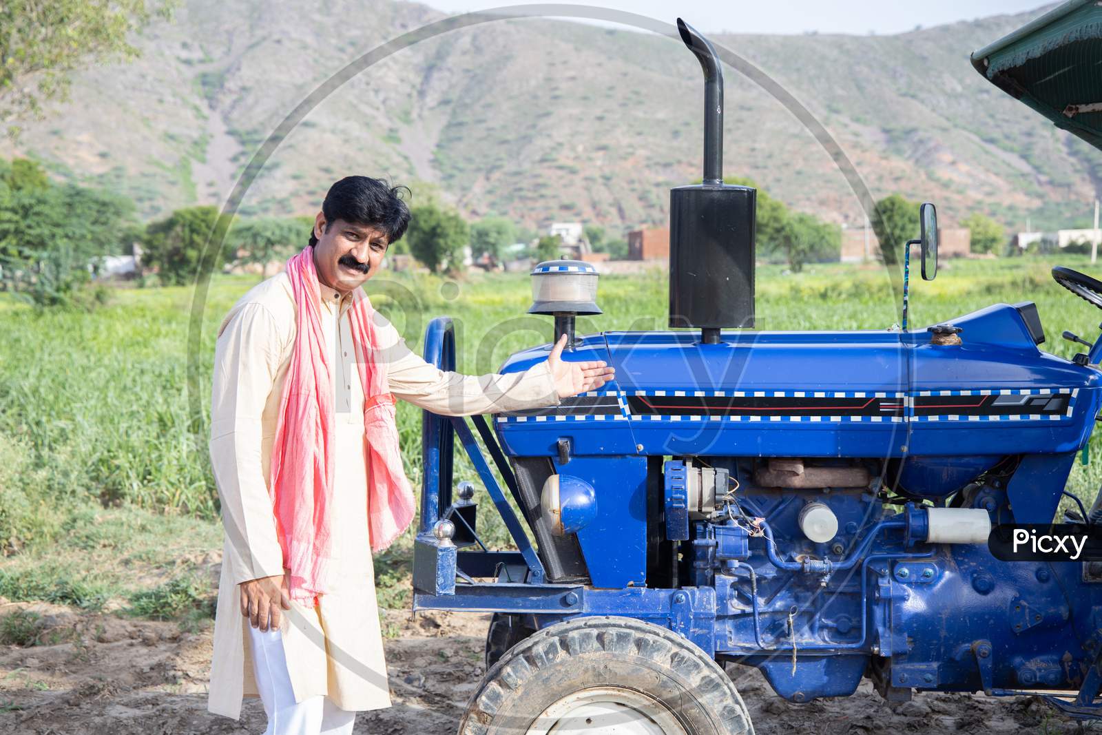 Young Happy Indian Farmer Showing His Blue Tractor Standing At Agriculture Field. Man Wearing Traditional Kurta With Mustache Smiling Looking At Camera. Rural India Concept.