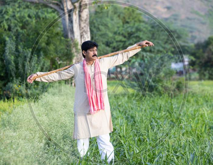 Indian Farmer Holding Wood Stick In Hand Standing In Agriculture Field Wearing Traditional Kurta Dress, Man With Mustache And Black Hair. Rural India Concept.