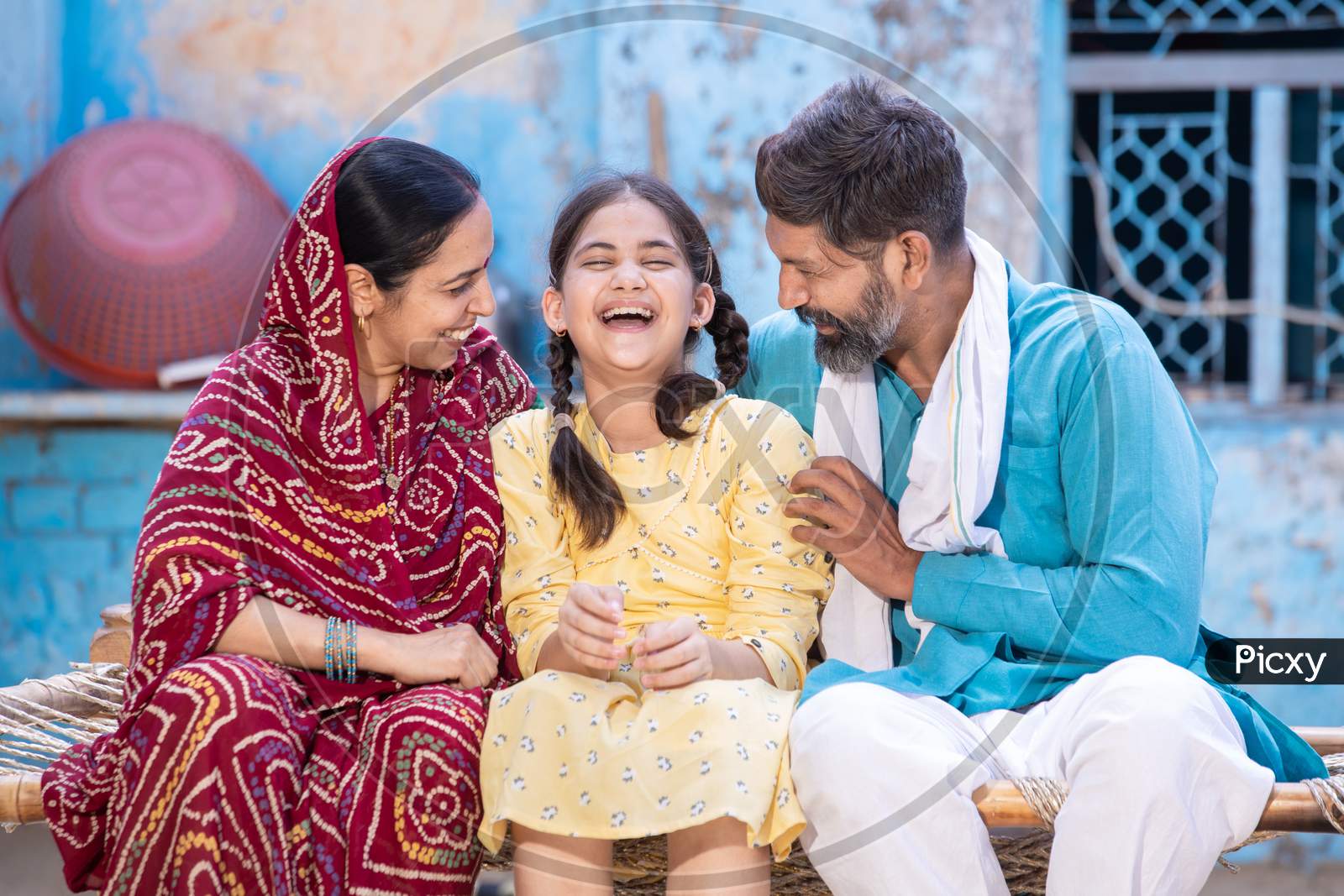Portrait Of Happy Rural Indian Family Laughing And Having A Good Time Together While Sitting Outside At Village House, Cheerful Little Daughter Enjoy With Her Father And Mother.Parent With Child