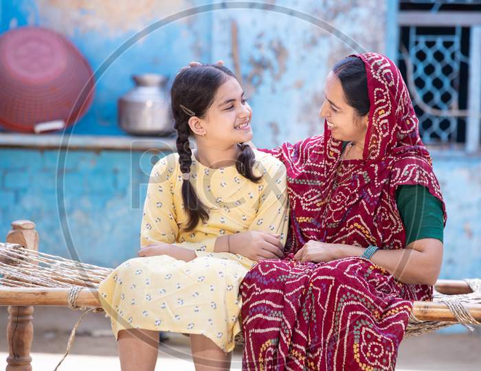 Smiling Young Indian Mother And Adorable Little Daughter Having A Good Time Together, Sitting On Traditional Bed, Cute Child Girl In Braided Hair And Mum In Red Sari Looking At Each Other.