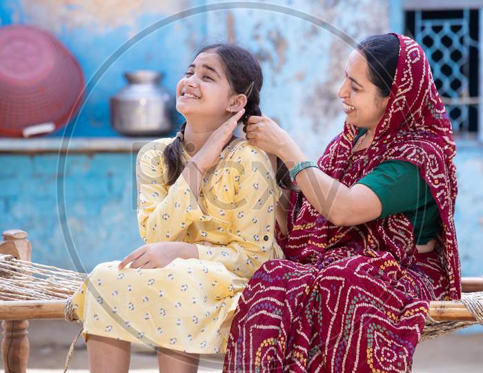 Rural Indian Mother Braids Hair Of Her Young Adorable Daughter, Cute Girl Feel Pain As Mum Getting Her Ready Sitting On Traditional Bed At Village Home. Both Spending Time Together.
