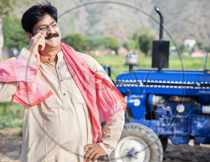 Young Happy Indian Farmer Touching His Mustache Standing At Agriculture Field With Blue Tractor Behind Him. Man Wearing Traditional Kurta Feel Proud. Rural India Concept.