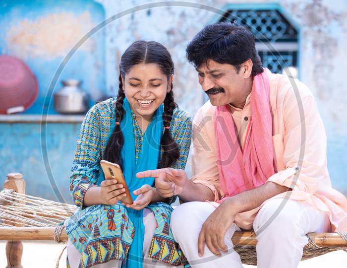 Happy Indian Father And Young Daughter Using Smartphone While Sitting On Traditional Bed Outside Their House, Happy Rural Family, Man Point Finger At Mobile Screen. Video Call, Learning Technology