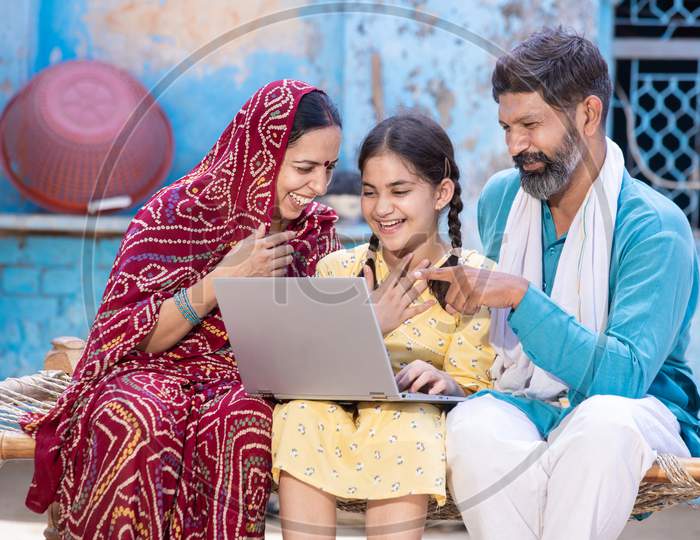 Happy Indian Little Girl Child Using Laptop While Sitting With Her Father And Mother. Village Parents Motivate Support Their Daughter For Online Education. Digital India. Man Point Finger At Computer