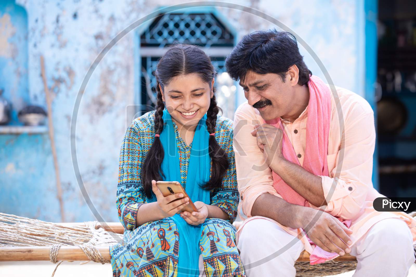 Happy Indian Father And Young Daughter Using Smartphone While Sitting On Traditional Bed , Happy Rural Family Watching At Mobile Screen. Internet Video Zoom Call, Digital India.