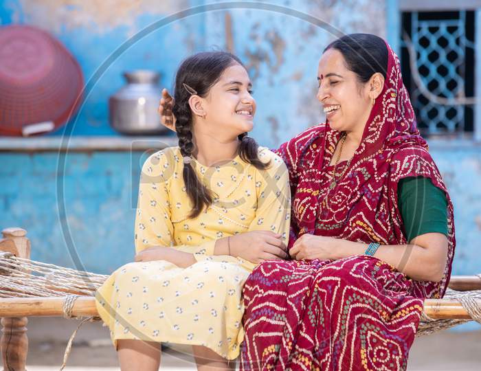 Smiling Young Indian Mother And Adorable Little Daughter Having A Good Time Together,Sitting On Traditional Bed, Cute Child Girl In Braided Hair And Mum In Red Sari Looking At Each Other
