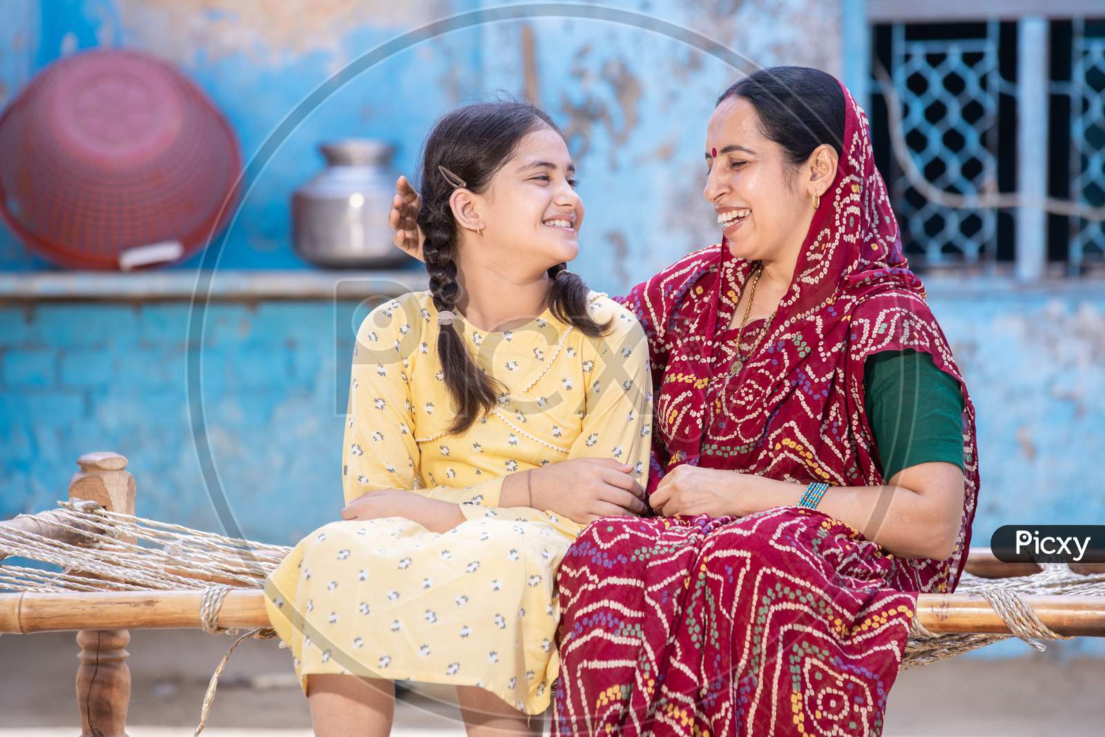 Smiling Young Indian Mother And Adorable Little Daughter Having A Good Time Together,Sitting On Traditional Bed, Cute Child Girl In Braided Hair And Mum In Red Sari Looking At Each Other