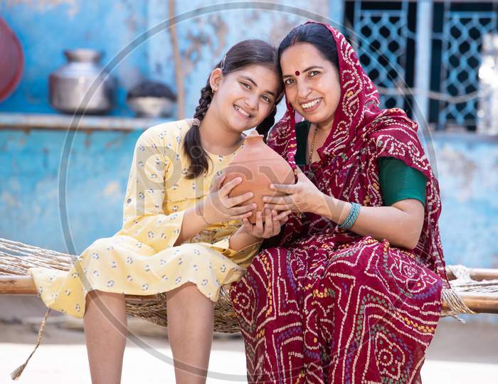 Portrait Of Happy Rural Indian Mother And Adorable Daughter Holding Piggy Bank While Sitting On Traditional Bed At Village Home, Cute Little Girl Collect Money, Saving And Investment Concept.
