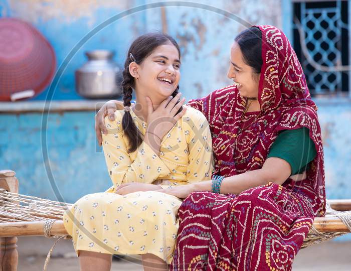 Cheerful Rural Indian Mother And Young Child Daughter Laughing Having A Good Time Together, Sitting On Traditional Bed, Cute Adorable Girl Kid In Braided Hair And Mum In Red Sari, Happy Parent Bonding