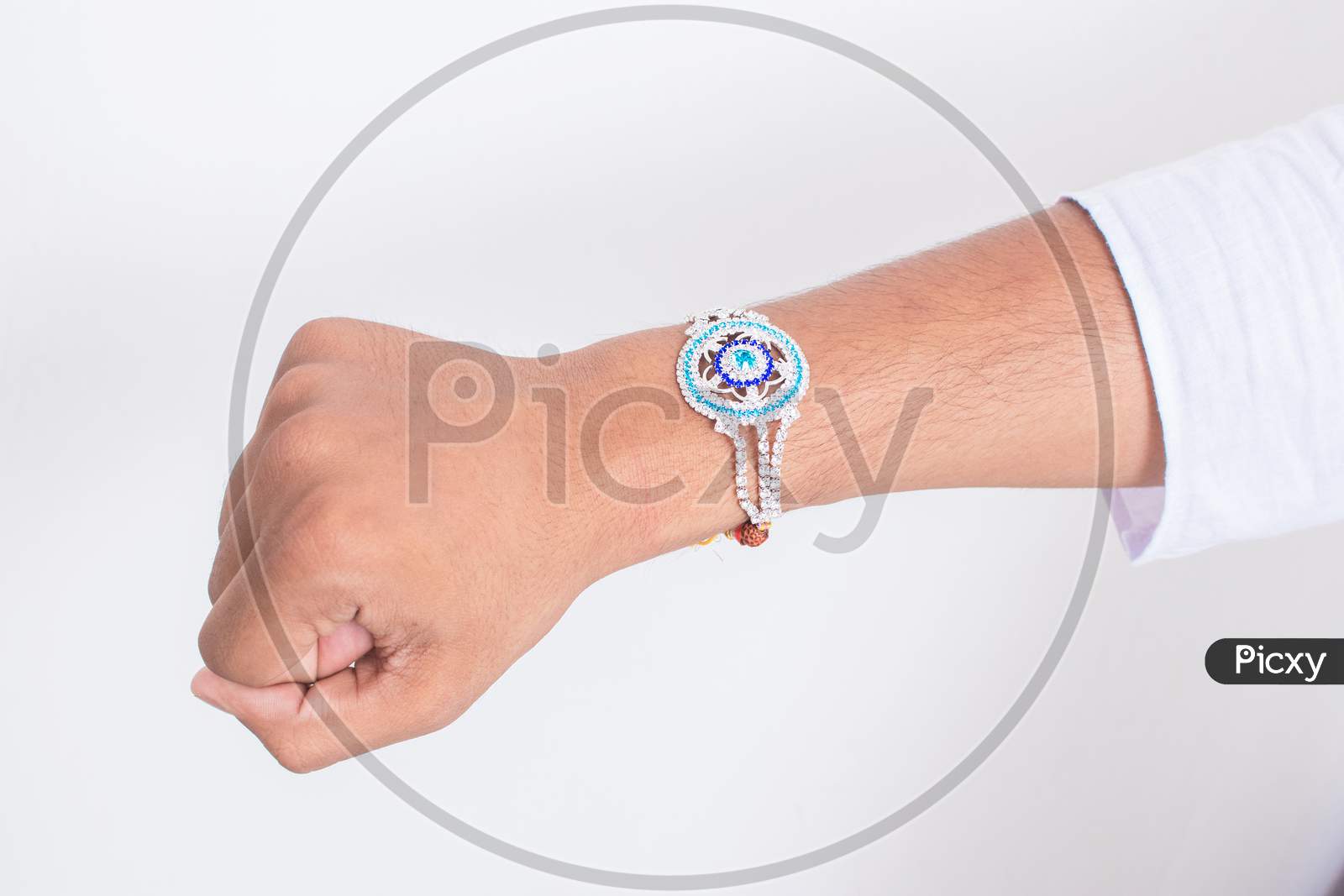 Hand Wearing Beautiful Blue Diamond Design Rakhi On The Occasion Of Raksha Bandhan Over White Background. Indian Festival Celebrated In India To Express Love And Bond Between Brother And Sister.